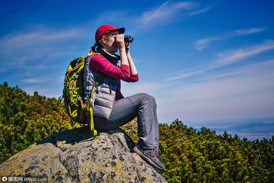 轻的女游客带着绿色的背包坐山顶上用望远镜看