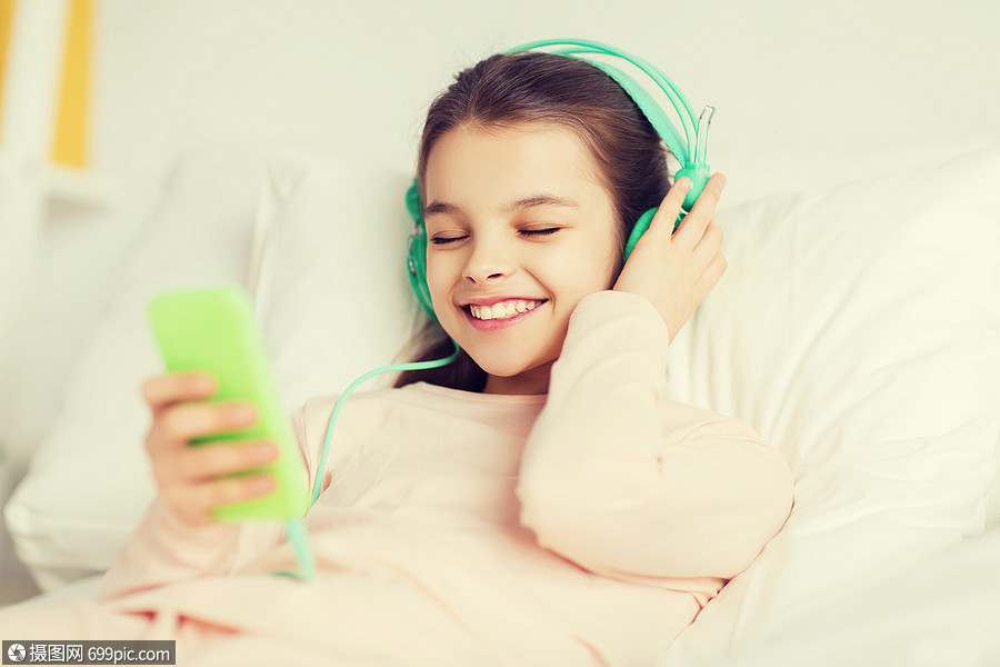 快乐的微笑女孩躺床上,用智能手机耳机听家里的音乐快乐的女孩躺床上